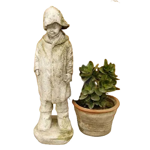 Statue of a child in a hooded coat with downcast gaze, old world European style, beside a potted plant with dense foliage.