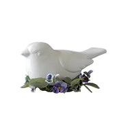White bird figurine in classical style, surrounded by ornate violet flowers and green leaves, evoking old world European charm.