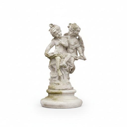 Pair of winged cherubs sitting on a round pedestal, one holding a scroll, with playful facial expressions.